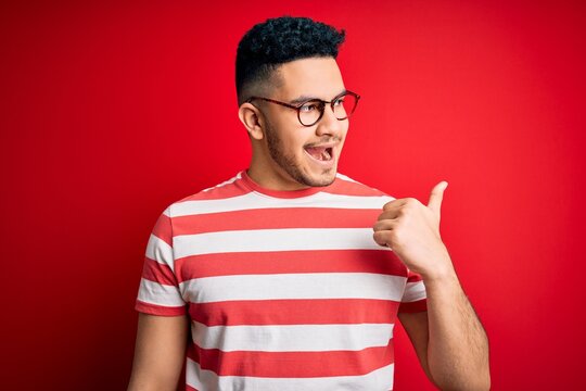 Young handsome man wearing casual striped t-shirt and glasses over isolated red background smiling with happy face looking and pointing to the side with thumb up.