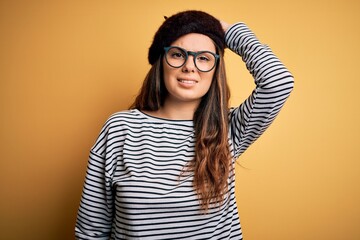 Young beautiful brunette woman wearing french beret and glasses over yellow background confuse and wonder about question. Uncertain with doubt, thinking with hand on head. Pensive concept.