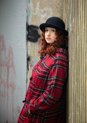 Portrait of a young woman with curly red hair, wearing a red tartan coat, set in a rough urban setting.