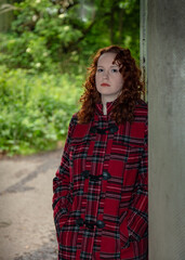 Portrait of a young woman with curly red hair, wearing a red tartan coat, set in a rough urban setting.