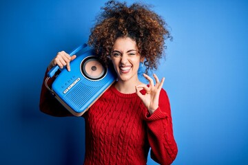 Obraz premium Young beautiful woman with curly hair and piercing listening to music using vintage radio doing ok sign with fingers, excellent symbol