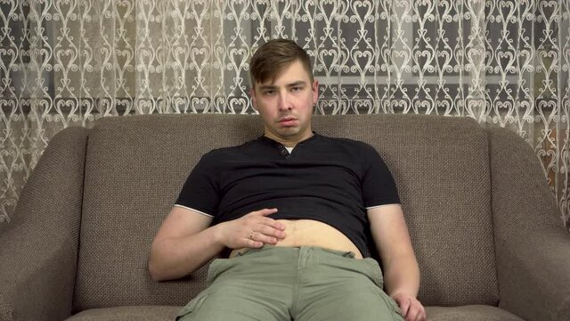 A young man touches his fat belly and then cries. The man on the couch slaps on his stomach. Gained excess weight in quarantine.