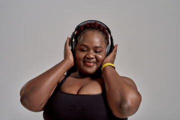 Plump, plus size african american woman in sportswear and headphones listening to music, standing in studio over grey background. Concept of sport, healthy lifestyle, body positive, equality