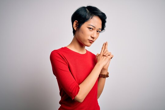 Young beautiful asian girl wearing casual red t-shirt standing over isolated white background Holding symbolic gun with hand gesture, playing killing shooting weapons, angry face