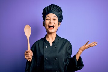 Fototapeta premium Young beautiful chinese chef woman wearing cooker uniform and hat holding wooden spoon very happy and excited, winner expression celebrating victory screaming with big smile and raised hands