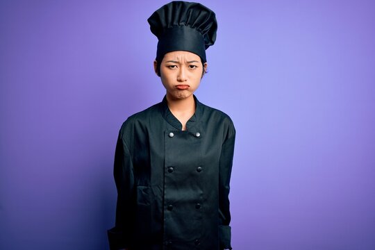 Young beautiful chinese chef woman wearing cooker uniform and hat over purple background depressed and worry for distress, crying angry and afraid. Sad expression.