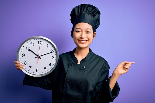 Young chinese chef woman wearing cooker uniform and hat holding clock doing countdown very happy pointing with hand and finger to the side