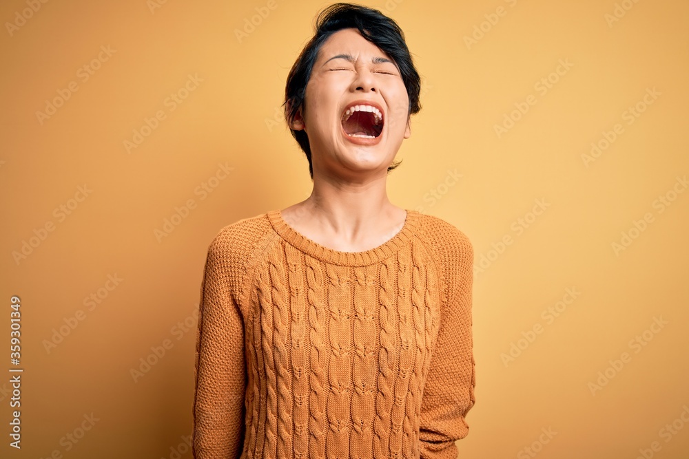 Poster Young beautiful asian girl wearing casual sweater and diadem standing over yellow background angry and mad screaming frustrated and furious, shouting with anger. Rage and aggressive concept.