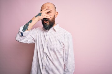 Handsome bald man with beard wearing elegant shirt over isolated pink background peeking in shock covering face and eyes with hand, looking through fingers with embarrassed expression.