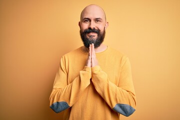 Handsome bald man with beard wearing casual sweater standing over yellow background praying with hands together asking for forgiveness smiling confident.