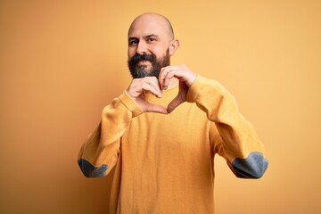 Handsome bald man with beard wearing casual sweater standing over yellow background smiling in love doing heart symbol shape with hands. Romantic concept.