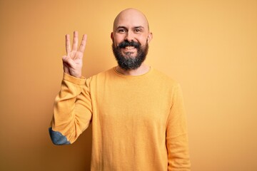 Handsome bald man with beard wearing casual sweater standing over yellow background showing and pointing up with fingers number three while smiling confident and happy.