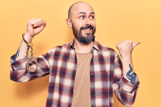 Young handsome man wearing prisoner handcuffs pointing thumb up to the side smiling happy with open mouth