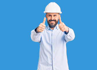 Young handsome man wearing architect hardhat approving doing positive gesture with hand, thumbs up smiling and happy for success. winner gesture.