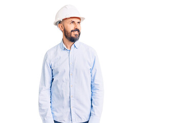Young handsome man wearing architect hardhat looking away to side with smile on face, natural expression. laughing confident.