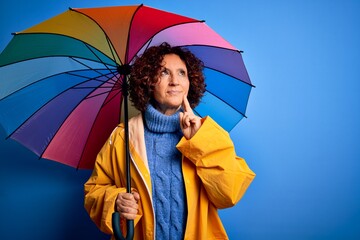 Middle age curly hair woman wearing rain coat holding colorful umbrella over blue background serious face thinking about question, very confused idea