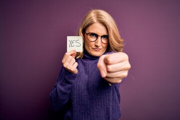 Middle age beautiful blonde woman wearing glasses holding reminder paper with yes message annoyed...