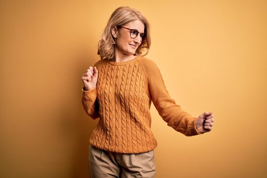 Middle age beautiful blonde woman wearing casual sweater and glasses over yellow background Dancing happy and cheerful, smiling moving casual and confident listening to music