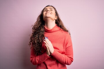 Young beautiful woman with curly hair wearing turtleneck sweater over pink background begging and...