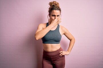 Young beautiful blonde sportswoman doing sport wearing sportswear over pink background smelling something stinky and disgusting, intolerable smell, holding breath with fingers on nose. Bad smell