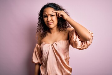 Young beautiful woman with curly hair wearing casual t-shirt standing over pink background pointing...