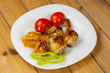 Tasty and crispy fried chicken legs, fried potatoes, tomatoes, bell pepper in a white plate on a wooden background.