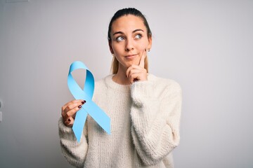 Young beautiful blonde woman holding blue cancer ribbon over isolated white background serious face thinking about question, very confused idea