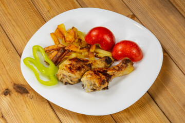 Tasty and crispy fried chicken legs, fried potatoes, tomatoes, bell pepper in a white plate on a wooden background.