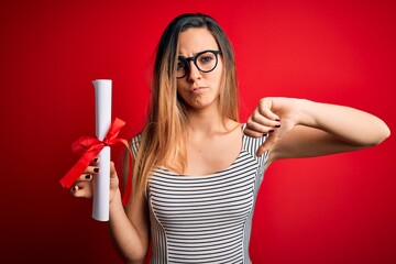 Young blonde woman with blue eyes wearing glasses holding graduated degree diploma with angry face, negative sign showing dislike with thumbs down, rejection concept