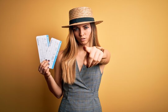 Young blonde tourist woman with blue eyes on vacation wearing hat holding boarding pass pointing with finger to the camera and to you, hand sign, positive and confident gesture from the front