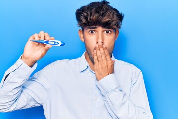 Young hispanic man holding thermometer covering mouth with hand, shocked and afraid for mistake....