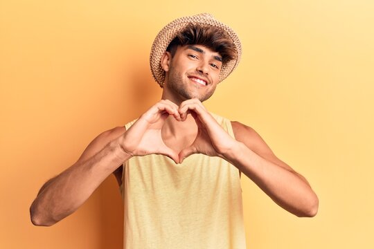 Young hispanic man wearing summer hat smiling in love doing heart symbol shape with hands. romantic concept.