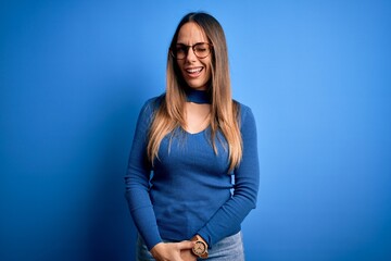 Young beautiful blonde woman with blue eyes wearing glasses standing over blue background winking...