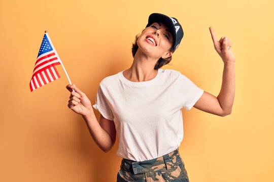Young beautiful woman wearing usa cap holding united states flag smiling with an idea or question pointing finger with happy face, number one