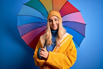Young beautiful blonde woman wearing raincoat for rainy weather holding colorful umbrella with a...