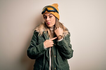 Young brunette skier woman wearing snow clothes and ski goggles over white background In hurry pointing to watch time, impatience, looking at the camera with relaxed expression