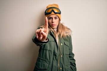Young brunette skier woman wearing snow clothes and ski goggles over white background Pointing with finger up and angry expression, showing no gesture