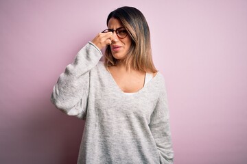 Young beautiful brunette woman wearing casual sweater and glasses over pink background smelling something stinky and disgusting, intolerable smell, holding breath with fingers on nose. Bad smell