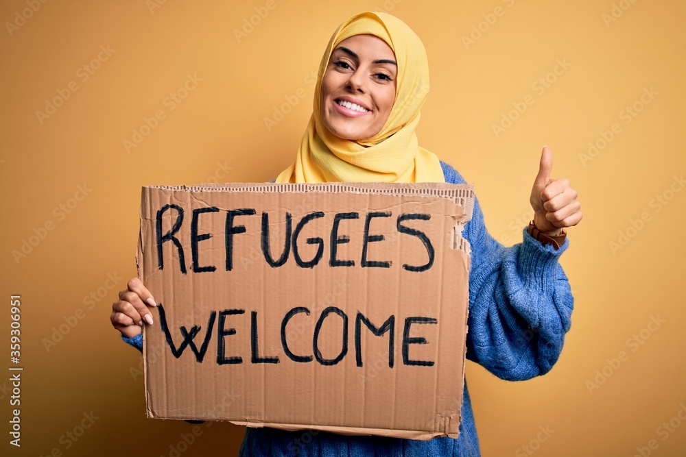 Sticker Beautiful arab woman wearing islamic hijab holding banner with refugees welcome message happy with big smile doing ok sign, thumb up with fingers, excellent sign