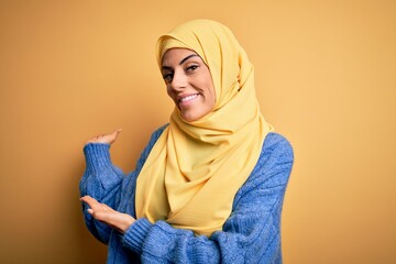 Obraz premium Young beautiful brunette muslim woman wearing arab hijab over isolated yellow background Inviting to enter smiling natural with open hand