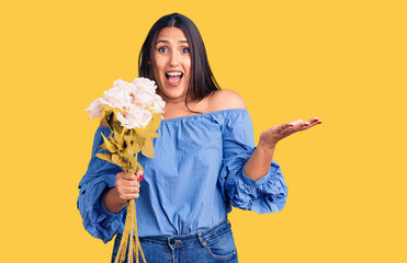 Young beautiful brunette woman holding bouquet celebrating victory with happy smile and winner expression with raised hands