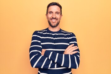 Young handsome man wearing casual striped sweater standing over isolated yellow background happy face smiling with crossed arms looking at the camera. Positive person.
