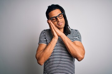 Young handsome african american man with dreadlocks wearing striped t-shirt and glasses sleeping tired dreaming and posing with hands together while smiling with closed eyes.