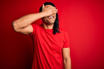 Young handsome african american afro man with dreadlocks wearing red casual t-shirt smiling and laughing with hand on face covering eyes for surprise. Blind concept.