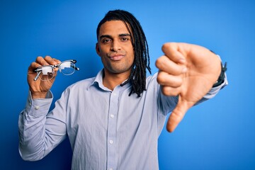 Young african american afro man with dreadlocks holding glasses over blue background with angry...