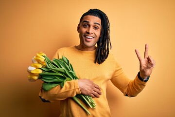 Young african american afro romantic man with dreadlocks holding bouquet of yellow tulips smiling looking to the camera showing fingers doing victory sign. Number two.