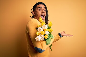 Young african american afro romantic man with dreadlocks holding bouquet of yellow tulips pointing aside with hands open palms showing copy space, presenting advertisement smiling excited happy