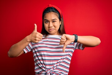 Young beautiful asian girl wearing casual striped t-shirt over isolated red background Doing thumbs up and down, disagreement and agreement expression. Crazy conflict