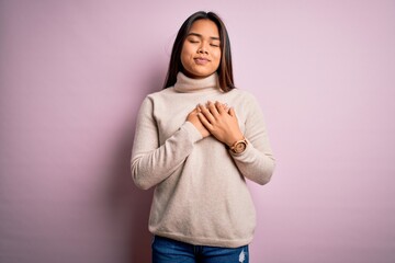 Young beautiful asian girl wearing casual turtleneck sweater over isolated pink background smiling with hands on chest with closed eyes and grateful gesture on face. Health concept.