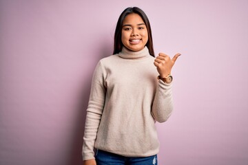 Young beautiful asian girl wearing casual turtleneck sweater over isolated pink background smiling with happy face looking and pointing to the side with thumb up.